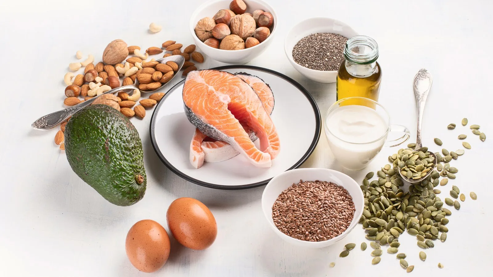 A spread of omega-3 food sources on a white surface: a salmon steak, avocado, nuts, seeds, eggs and a small bottle of oil.