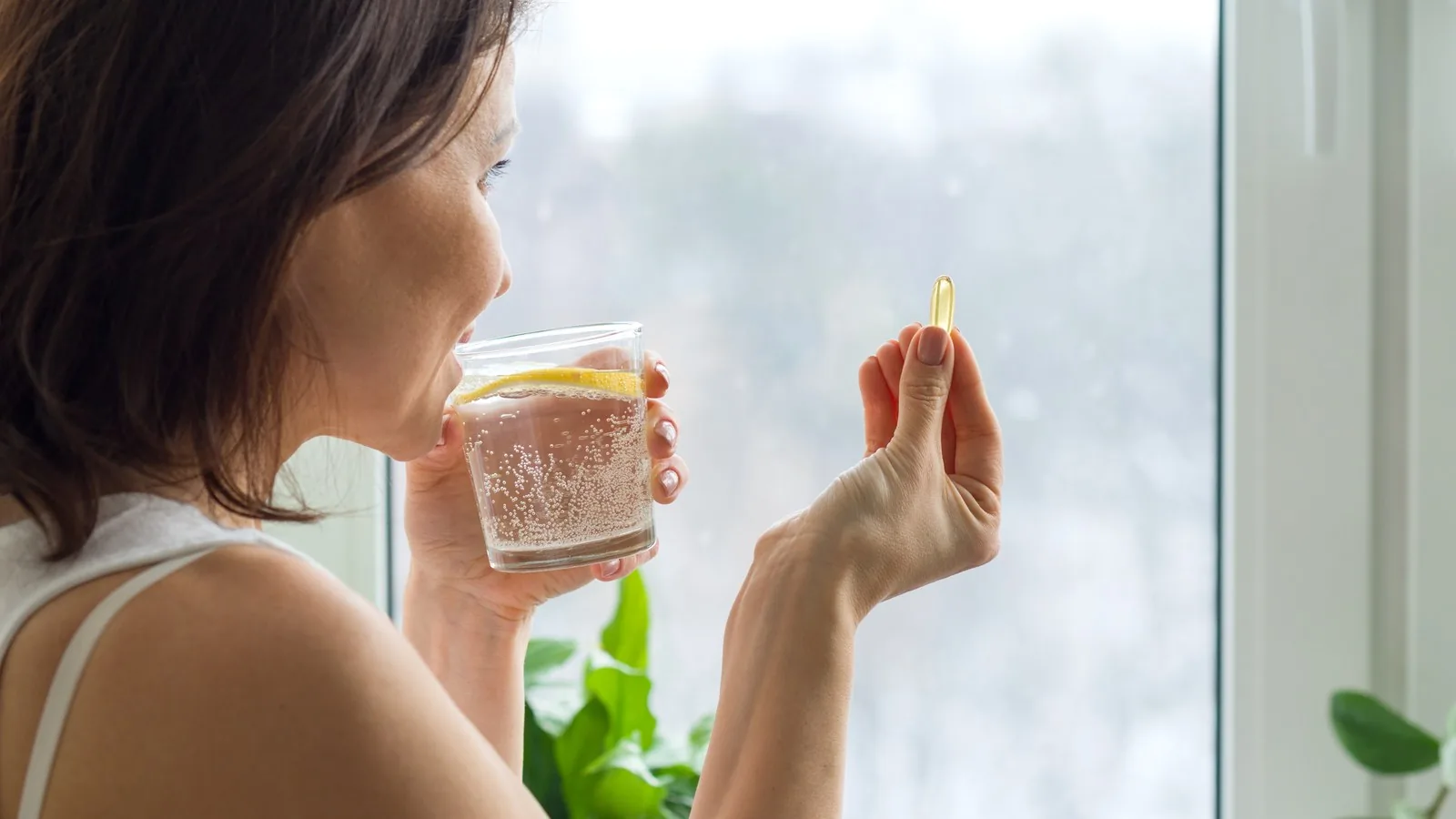 A woman at a bright window holding a single golden softgel in one hand and a glass of water in the other.